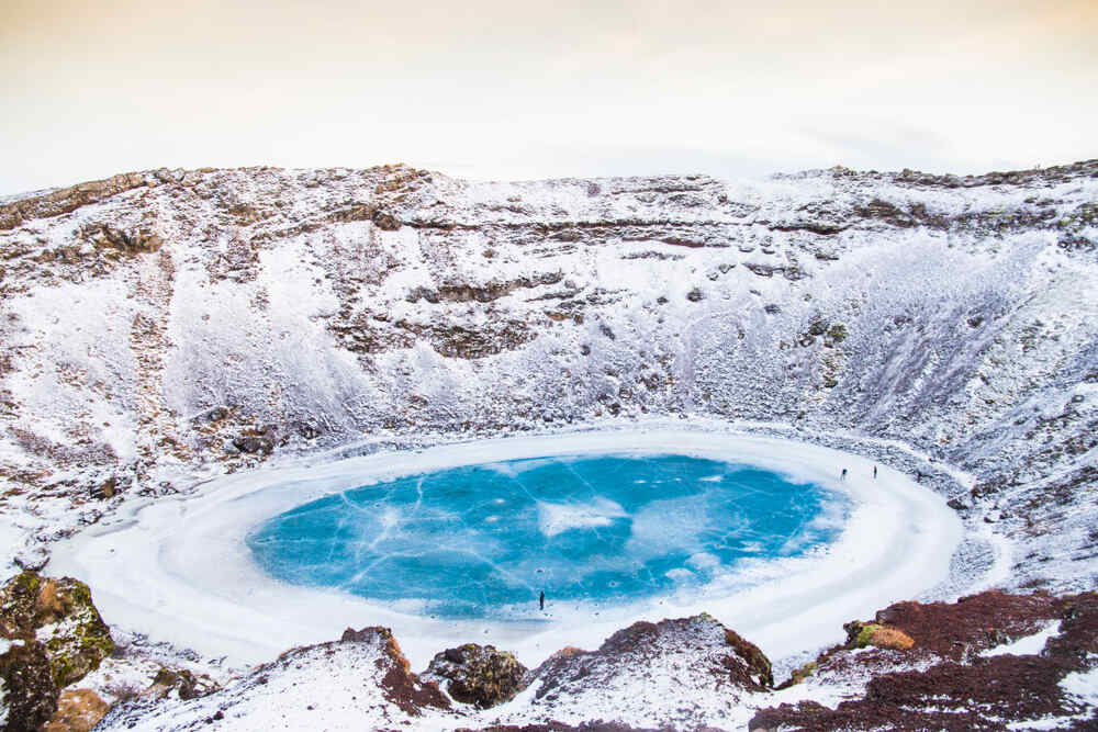 Le cratère Kerið recouvert de neige avec un lac glacé bleu brillant en son centre, entouré de falaises enneigées.