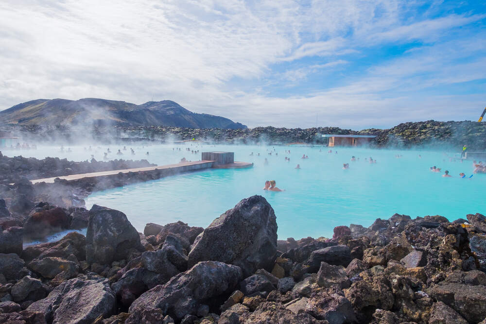 Le lagon bleu fumant entouré de roches volcaniques noires sous un ciel bleu clair.