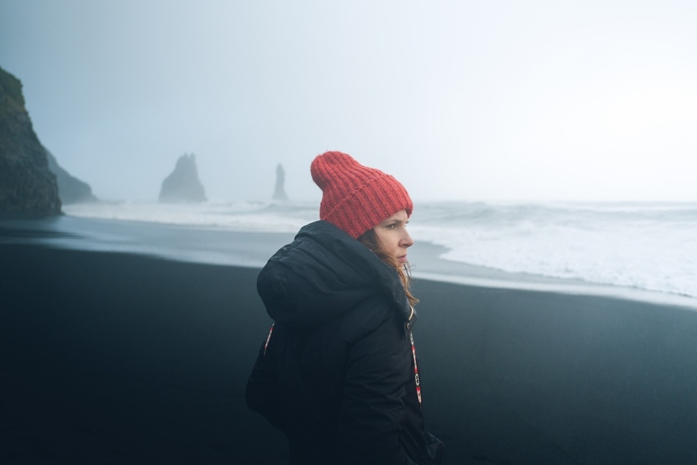 Une jeune femme portant un bonnet rouge marchant sur la plage de sable noir de Reynisfjara sous un ciel brumeux.