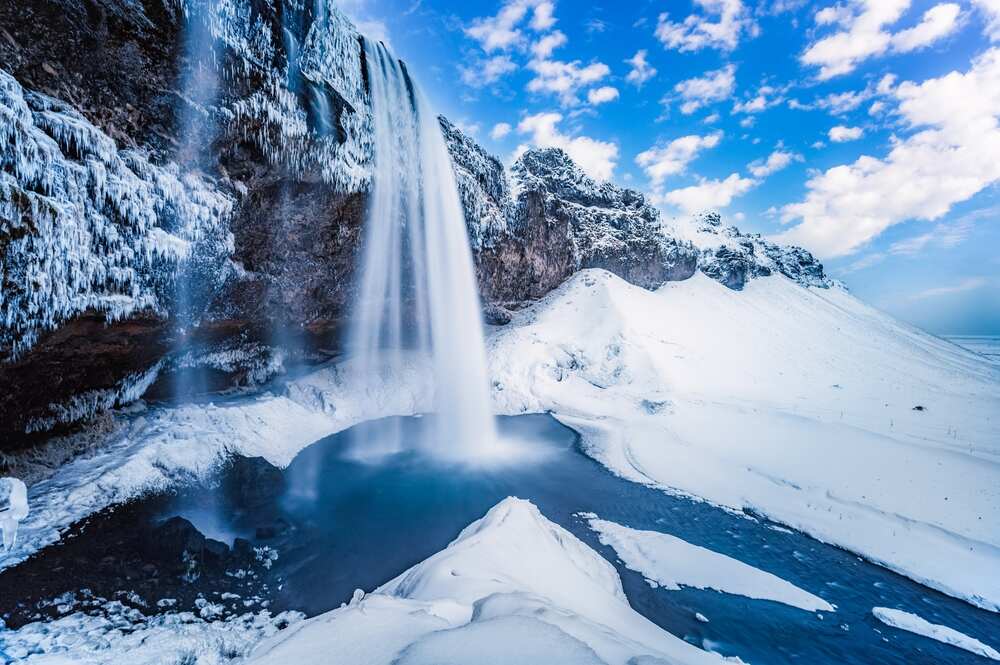 La cascade Seljalandsfoss en Islande en plein hiver, entourée de neige immaculée et de stalactites de glace, avec l'eau tombant en un rideau majestueux dans un bassin gelé.