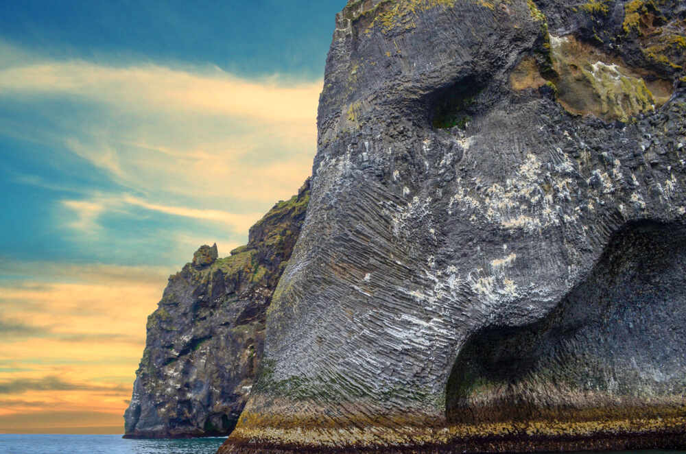 Une imposante falaise rocheuse sur l'île d'Heimaey, sculptée de manière à ressembler à une tête d'éléphant, se détache sur un ciel aux teintes de coucher de soleil.