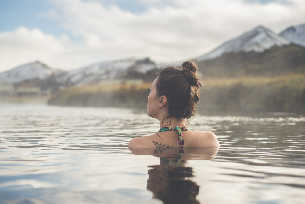 Bain relaxant dans une source chaude islandaise Une femme profitant des eaux d'une source chaude naturelle en Islande, avec un paysage montagneux en arrière-plan.
