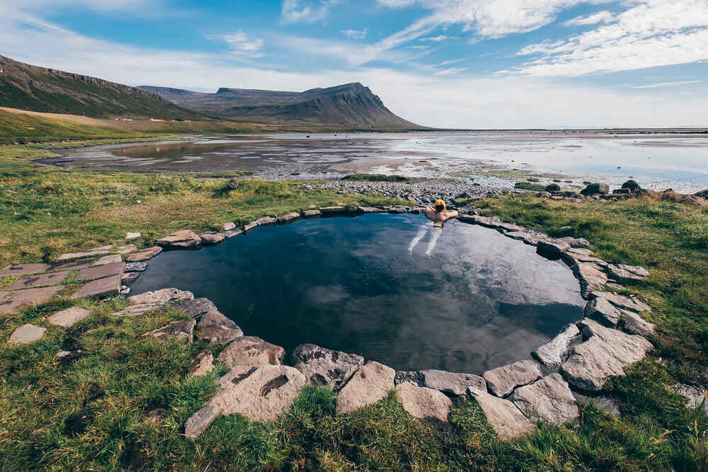 Source chaude isolée en Islande avec vue sur l'océan Une personne profitant d'une petite source chaude en plein air avec vue sur la mer et les montagnes environnantes.