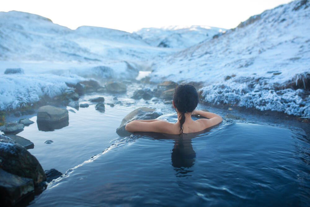 Bain chaud en hiver au cœur de la nature islandaise Une femme se relaxant dans une source chaude en plein hiver, entourée de paysages enneigés et de ruisseaux gelés.