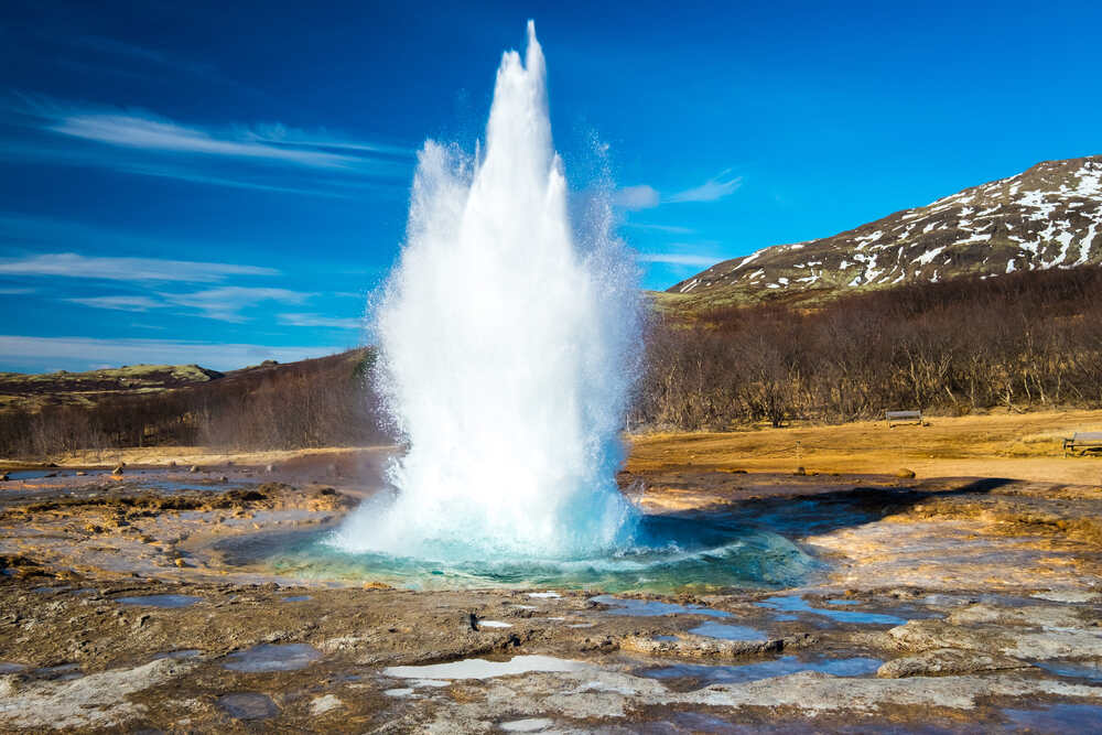 Geyser en éruption dans la région du Cercle d'Or en Islande Un geyser éclatant dans les airs avec une puissante colonne d'eau sous un ciel bleu clair.