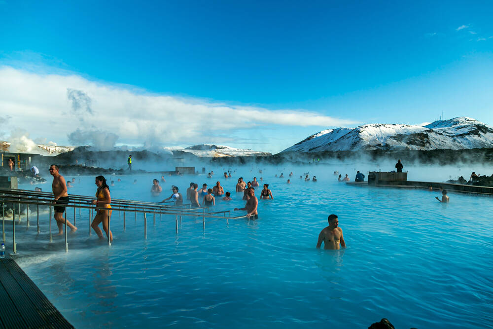 Détente à la lagune bleue d'Islande Des baigneurs profitant de la célèbre lagune bleue, entourés de vapeur et de montagnes enneigées sous un ciel bleu.