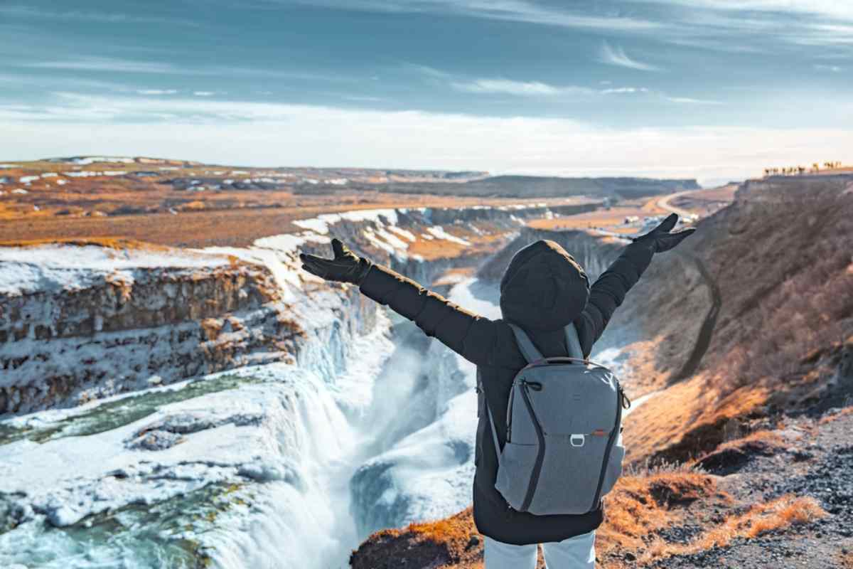 Une femme vêtue d’habits d’hiver lève les bras en croix en signe de joie après avoir regardé d’un point élevé la cascade Gullfoss complètement gelée.