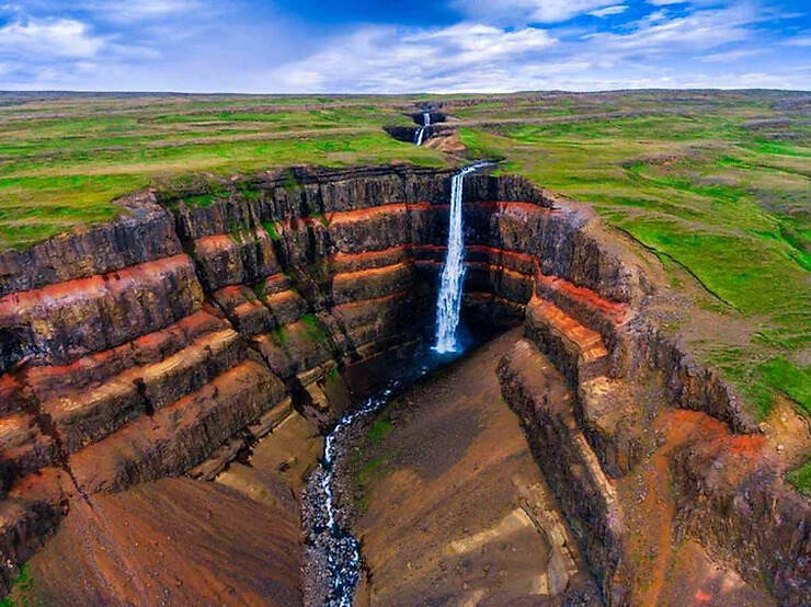 La maestosa cascata di Hengifoss, con le sue pareti di basalto stratificato e rosso, si tuffa in una gola profonda, immersa in un paesaggio verde islandese.