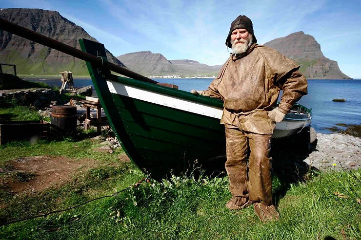 Uomo vestito con abiti tradizionali islandesi accanto a una barca storica su una costa pittoresca.