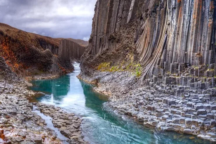 Vista di un canyon mozzafiato in Islanda, caratterizzato da alte colonne di basalto che si innalzano sopra un fiume color turchese, immerso in un'atmosfera drammatica con cielo nuvoloso.