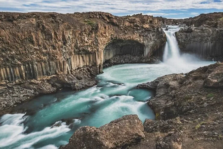 Cascata spettacolare circondata da formazioni uniche di colonne basaltiche nere. L'acqua cristallina si infrange con forza, creando un contrasto drammatico con il paesaggio vulcanico circostante.