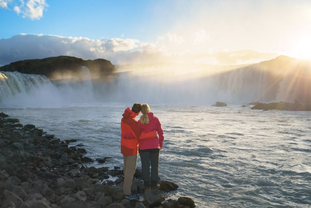 Una coppia si abbraccia di fronte alla spettacolare cascata Goðafoss, circondata da una luce dorata del tramonto.