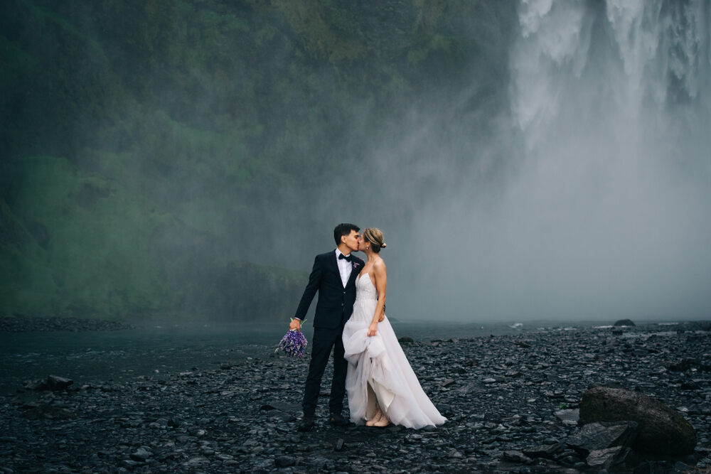 Una coppia vestita elegantemente celebra il proprio matrimonio di fronte alla cascata di Skógafoss, immersa in una cornice magica di acqua e natura.