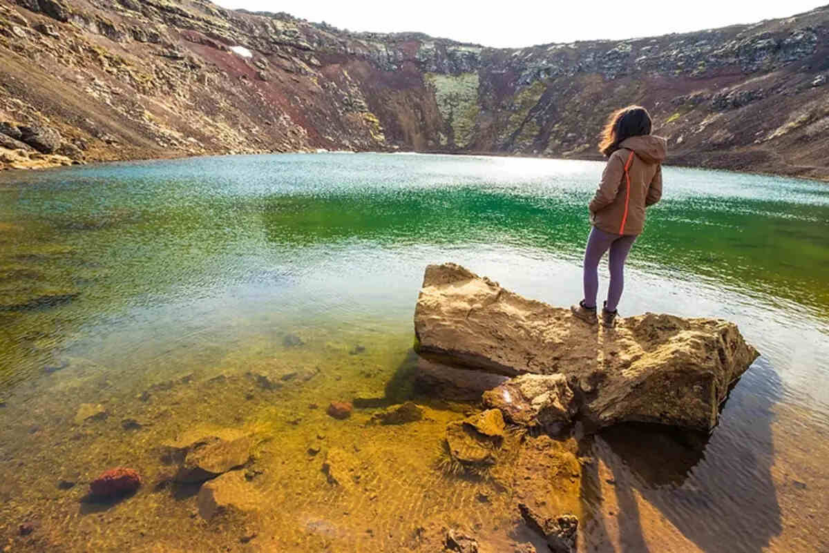 Una persona in piedi su una roccia osserva un lago color verde smeraldo all'interno del cratere di Kerið, circondato da pareti di roccia vulcanica.