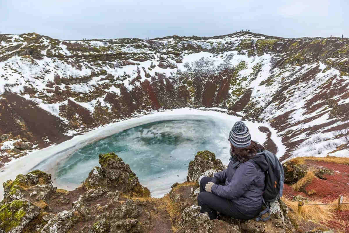 Una persona seduta su una roccia guarda un cratere vulcanico parzialmente ghiacciato, circondato da neve e paesaggi invernali.