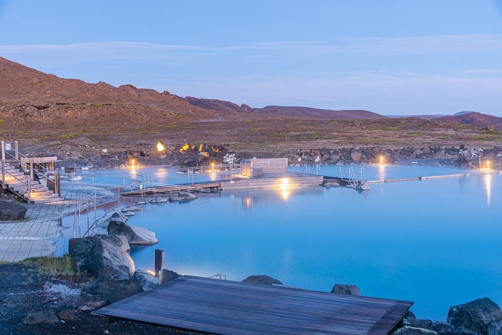 Una piscina termale all'aperto circondata da rocce vulcaniche nere, illuminata da luci soffuse. L'acqua è di un azzurro cristallino, che contrasta con l'ambientazione vulcanica circostante. Sullo sfondo, un paesaggio brullo e collinare.