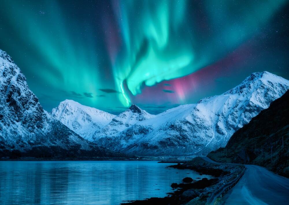 Un paesaggio mozzafiato con montagne innevate riflesse su un lago calmo. Il cielo notturno è animato da aurore boreali verdi e blu, creando un contrasto magico con le cime bianche.