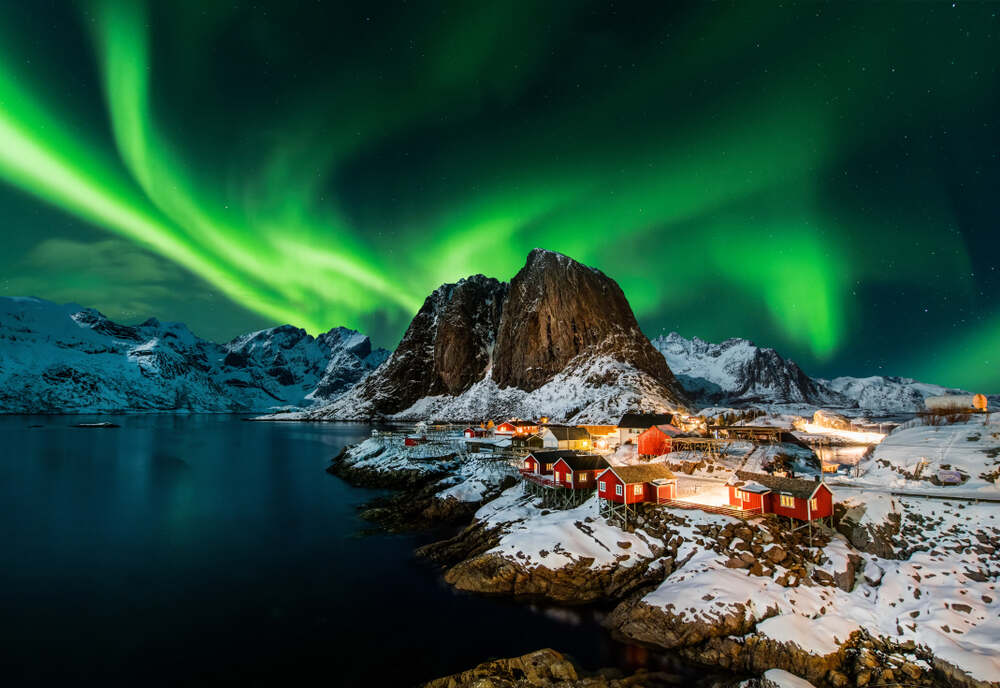 L'aurora boreale in Islanda Un villaggio costiero con case rosse e tetti innevati, illuminato dalle aurore boreali verdi e luminose che riempiono il cielo stellato sopra le montagne circostanti.