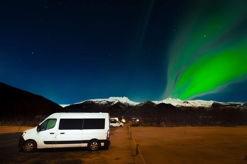 Aurora boreale islandese Un minivan bianco parcheggiato in un'area deserta e oscura. Sopra il veicolo, un'aurora boreale verde e brillante illumina il cielo notturno, creando una scena suggestiva con montagne sullo sfondo.