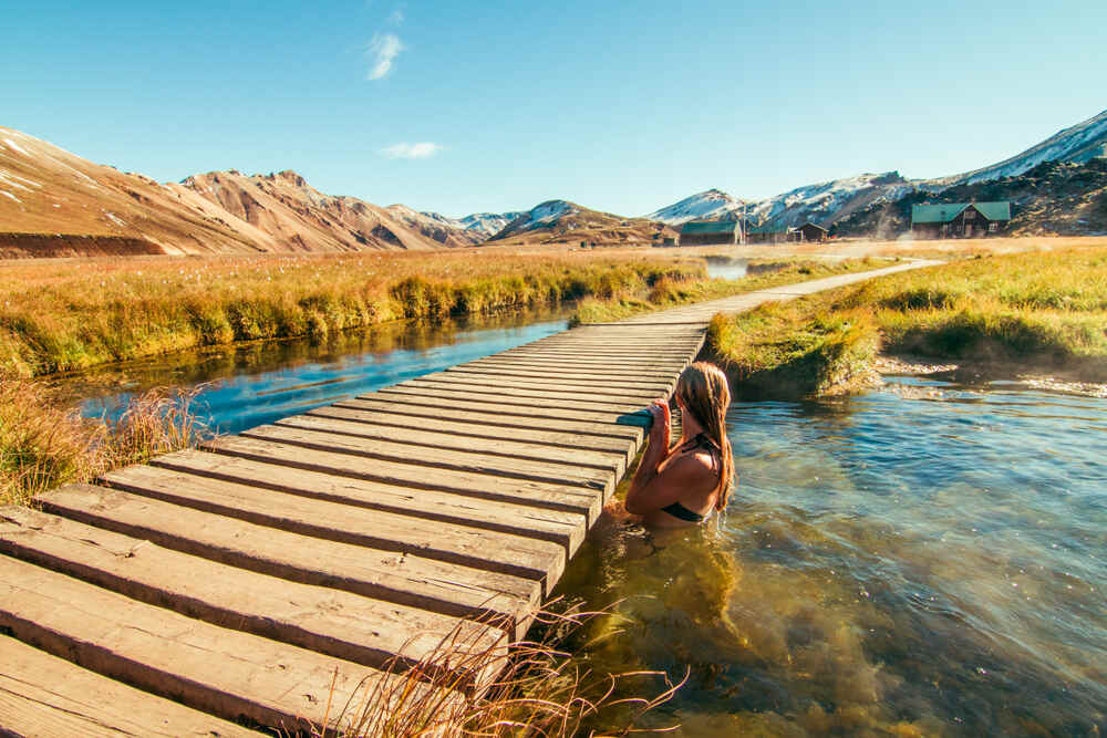 Una donna immersa in una sorgente termale naturale, con una passerella in legno e un paesaggio montano illuminato dal sole.