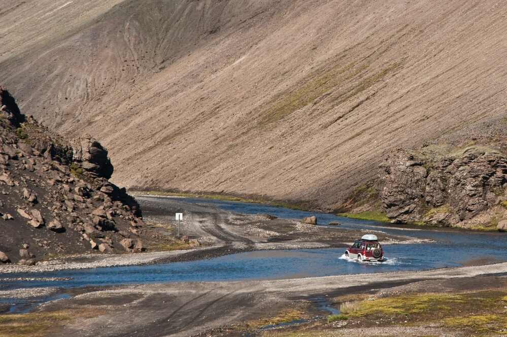 Un fuoristrada attraversa un piccolo fiume, circondato da un paesaggio montuoso con pendii rocciosi.