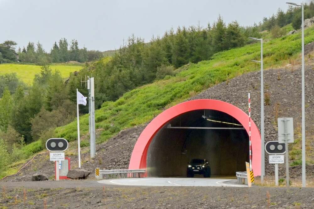 Pagamento del tunnel a pedaggio in Islanda Entrata di un tunnel caratterizzato da un arco rosso brillante e circondato da una vegetazione lussureggiante. Segnaletica di stop e altre indicazioni stradali presenti davanti all'entrata.