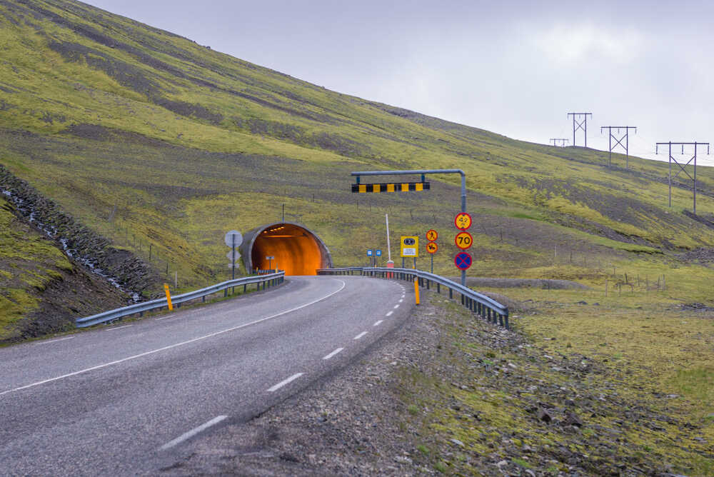 L'unica strada a pedaggio in Islanda Un tunnel arancione integrato in un paesaggio collinare verde. Segnali stradali gialli e rossi indicano limiti di velocità e altre restrizioni davanti all'ingresso del tunnel.