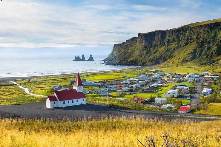 Una vista pittoresca del villaggio di Vík, con la sua chiesa in primo piano e l'Oceano Atlantico costeggiato da maestose scogliere sullo sfondo. Le famose formazioni rocciose di Reynisdrangar si stagliano all'orizzonte.