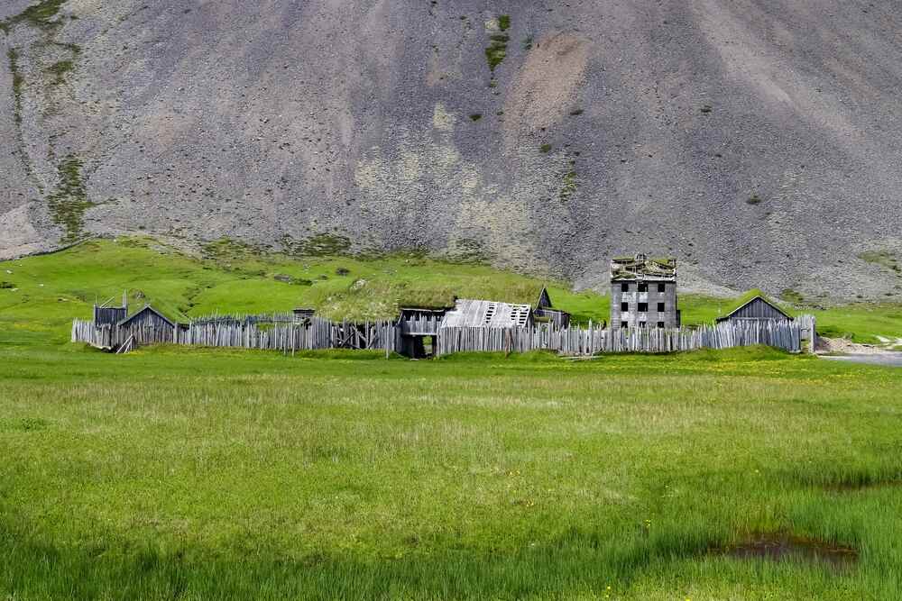 Una riproduzione di un antico villaggio vichingo con palizzate di legno e torrette, situato in una verde pianura ai piedi di una montagna grigia e ripida.