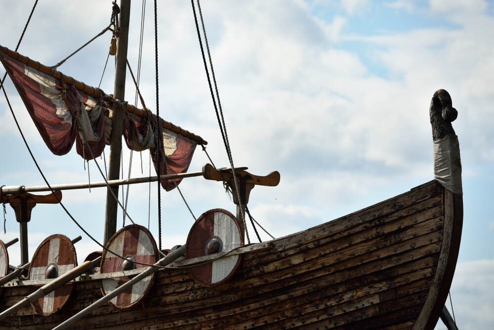 Dettaglio di una nave vichinga con scudi di legno colorati lungo i lati e vele piegate, ripresa con un cielo nuvoloso sullo sfondo.