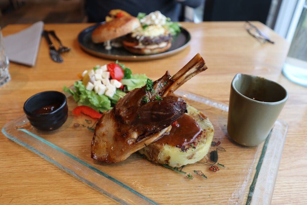 Un piatto di agnello arrosto servito su un vassoio di vetro, accompagnato da purè di patate e salsa, con un’insalata fresca di contorno e una tazza di sugo fumante.
