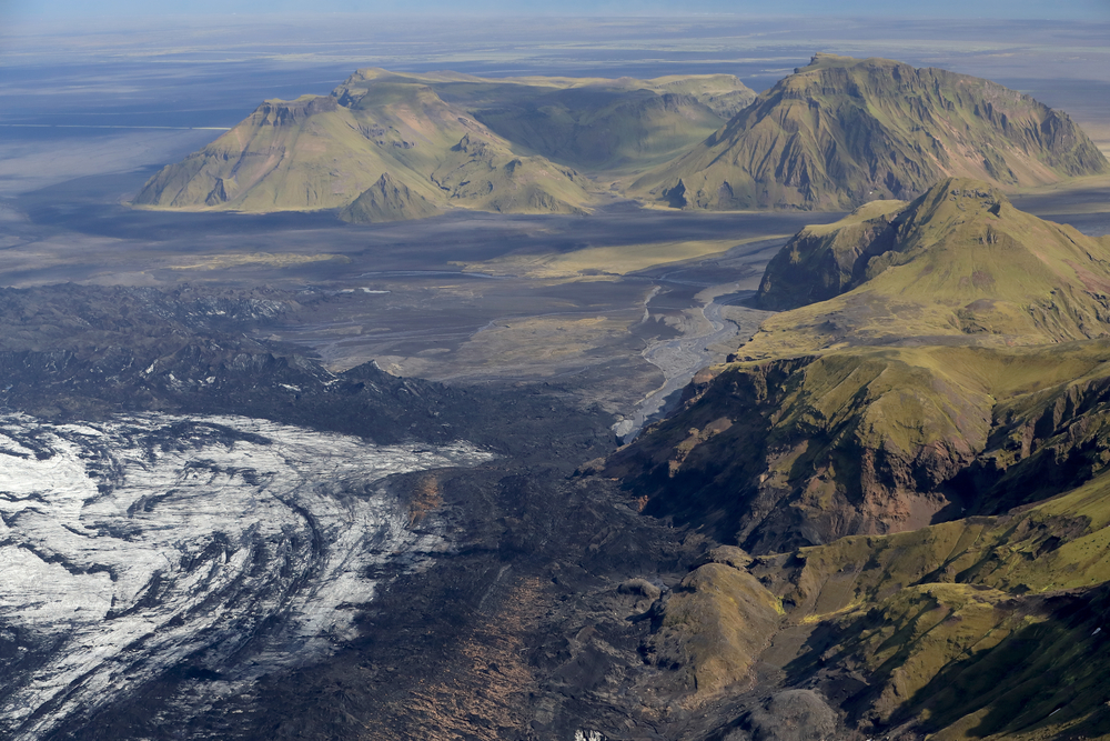 Aerial view of Katla volcano's black lava fields, glacial ice, and green ridges in Iceland’s Mýrdalsjökull area, showing stark natural contrasts.