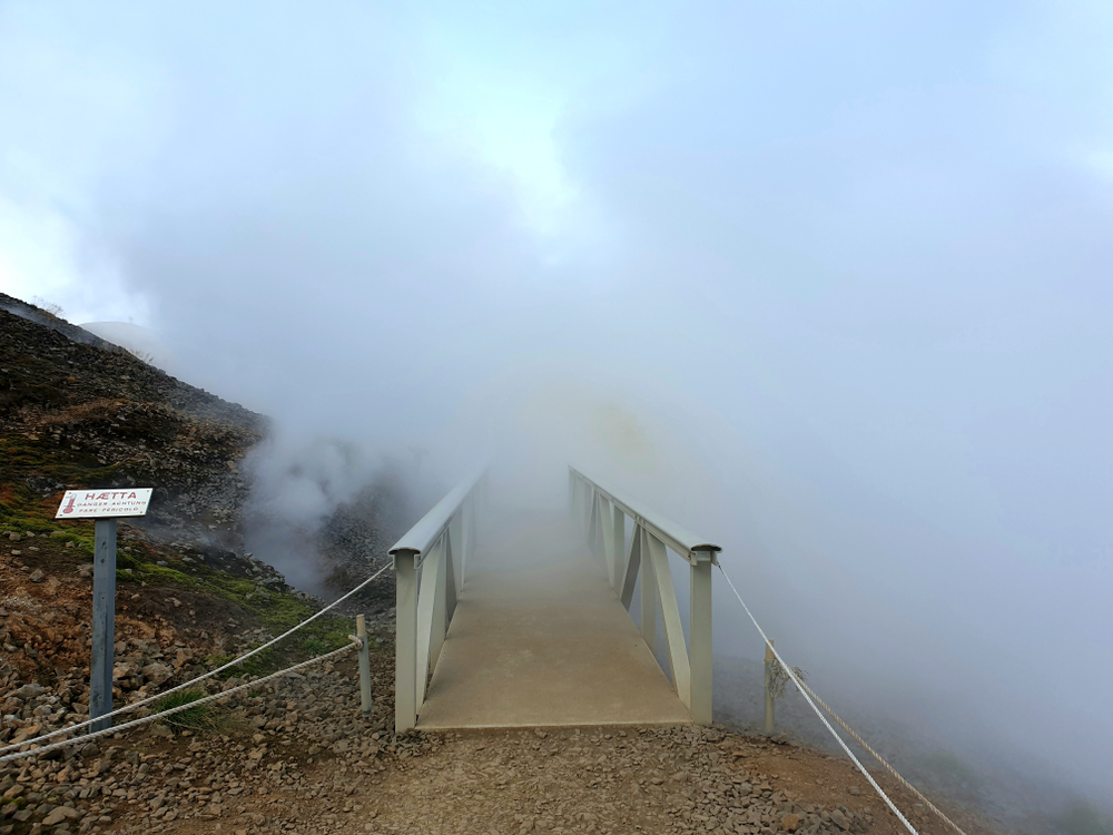 Attraversare il Vapore: Hverir, il Paesaggio Estremo delle Terre Geotermiche Islandesi Ponte immerso nel vapore geotermico nell’area termale di Hverir, Islanda, con cartello di pericolo che avverte delle alte temperature.