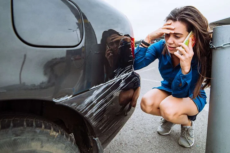 Una donna preoccupata inginocchiata accanto a un’auto a noleggio nera graffiata, al telefono mentre osserva i danni.